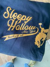 Close up detail photo of the Sleepy Hollow Headless Horseman baseball and horseman logo in orange on a folded black hooded sweatshirt.